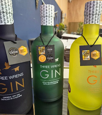 Three bottles of Three Wrens Gin on display, including original dry gin and flavoured varieties, with award badges on the labels, photographed at the Cheshire East distillery.