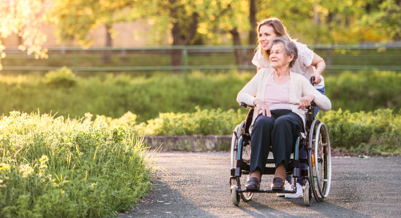 GettyImages-960150966 Elderly woman in wheelchair with an adult personal assistant outside in nature 570 x 310