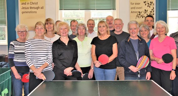 A group of 15 people stood in front of a table tennis table, some holding table tennis bats